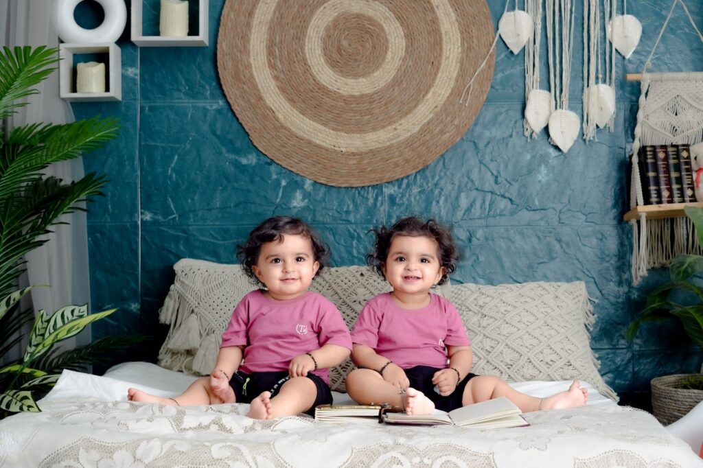 Cute twin toddlers in pink shirts sitting on a bed, surrounded by indoor decor.
