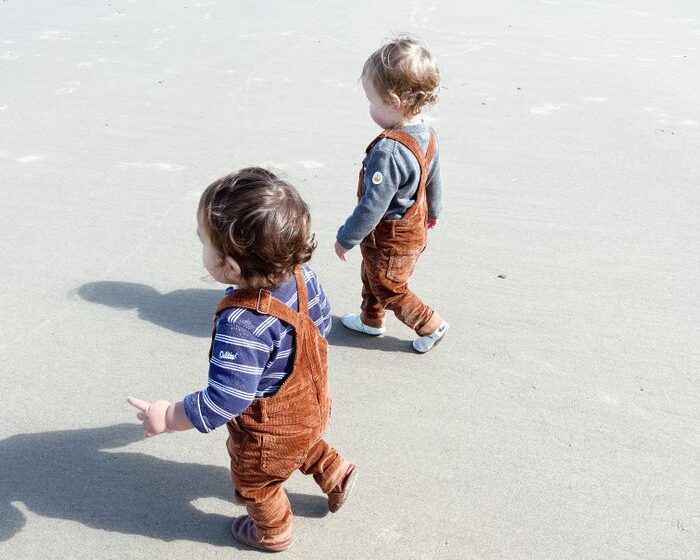 Two toddlers in orange overalls walking on a sandy beach, showcasing friendship and exploration.