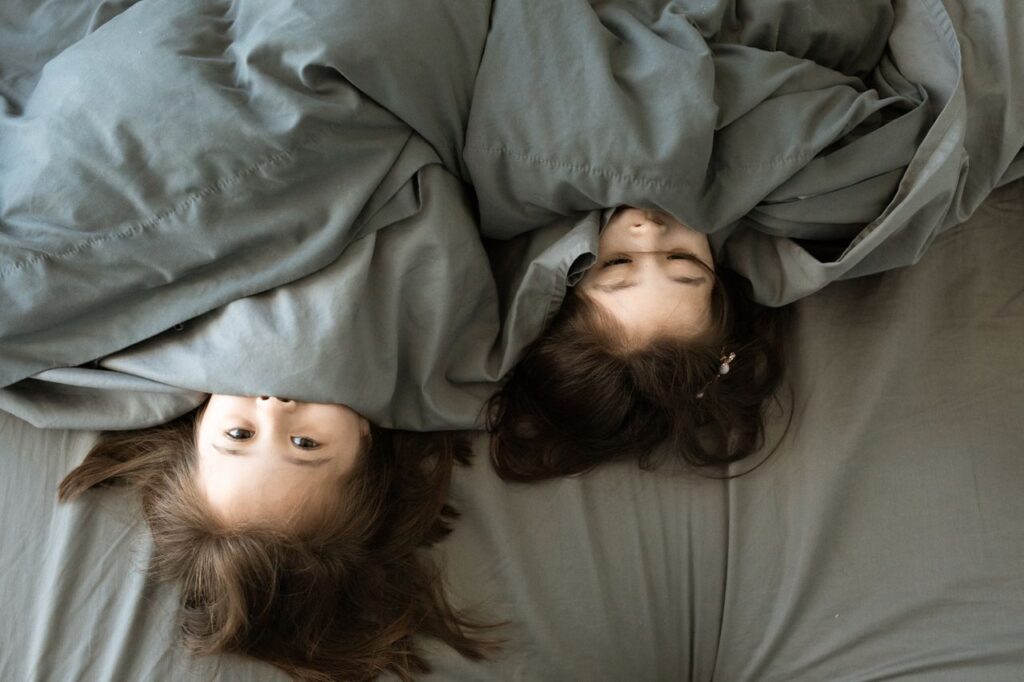 Two young girls playfully hiding under a grey blanket in bed, showing sweet togetherness.
