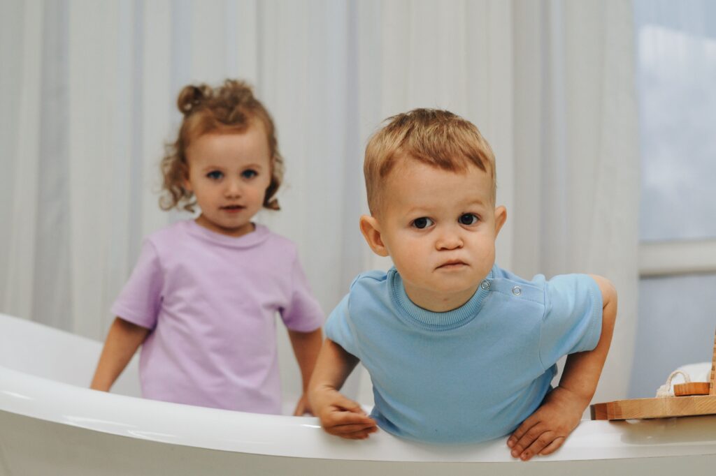 Two twin toddlers standing near a window — one wearing a pink shirt and the other in blue — looking curious and expressive in a softly lit room.