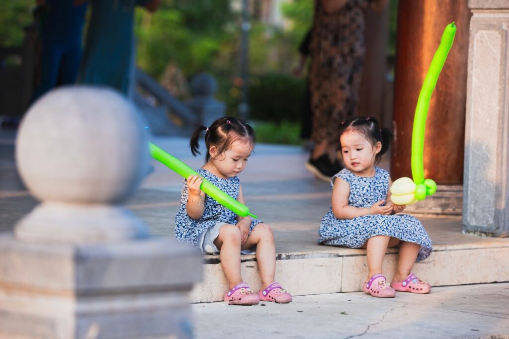 Two young girls sitting on steps with balloon swords, wearing matching dresses.