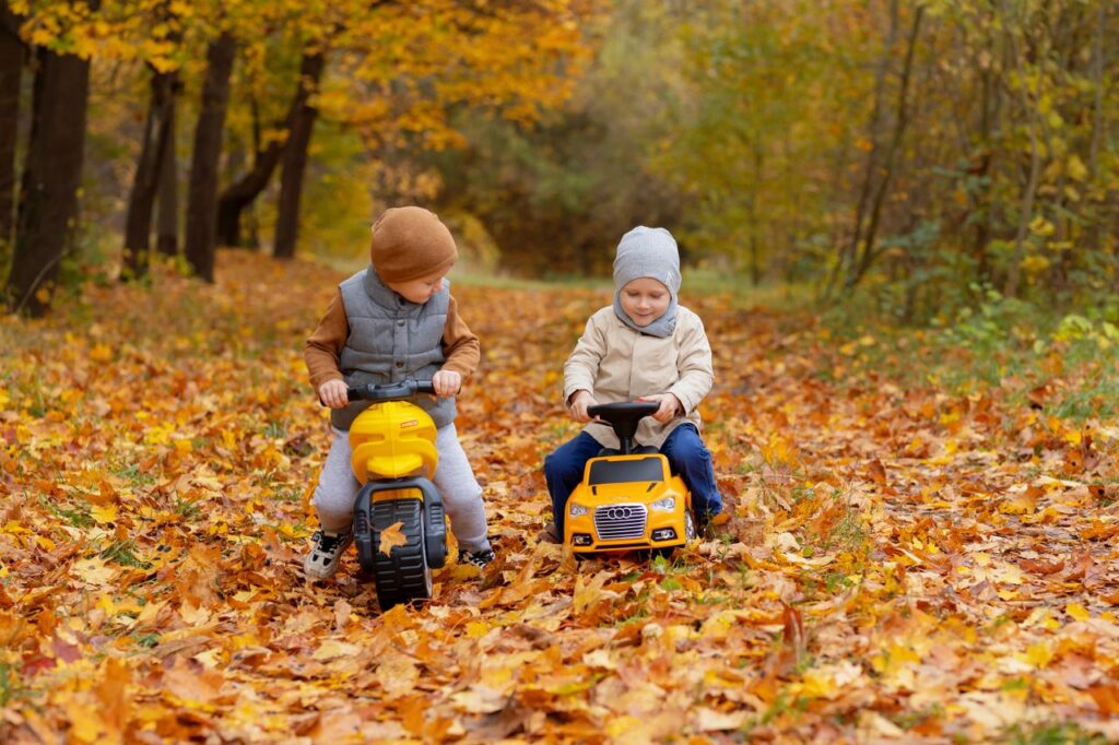 Two children playing with toy vehicles on a leaf-covered path during fall.